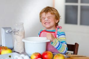 Cute little happy blond preschool kid boy baking apple cake and muffins in domestic kitchen. Funny lovely healthy child having fun with working with mixer, flour, eggs, fruits. Little helper indoors.